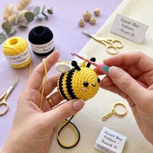 以下が含まれることがあります： 白い羽の黄色と黒のかぎ針編みの蜂が、ピンクのかぎ針で作業されています。 蜂は笑顔です。 黄色と黒の糸、はさみ、キーホルダーも表示されます。 「Kawaii Bee Pattern - Round 14」というテキストが小さなカードに書かれています。