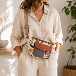 Peut inclure: Un sac banane en toile beige avec un imprimé botanique abstrait multicolore dans des tons bleu, vert et terre cuite. Le sac comprend une poche avant unie couleur terre cuite avec fermeture à rabat, une fermeture éclair en laiton et une sangle beige réglable.