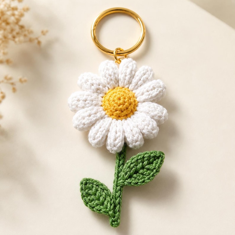May include: A handmade daisy keychain featuring a white crocheted flower with a yellow center and green stem and leaves. The keychain has a gold-colored ring for attaching to keys or bags. The daisy is a charming, floral accessory.