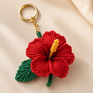 May include: A handmade red hibiscus flower keychain with a gold-colored ring. The flower has a yellow center and a green stem and leaf. The keychain is made of yarn and is a decorative accessory.