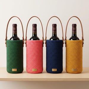 Könnte beinhalten: Vier Weinflaschenhalter in Grün, Rosa, Marineblau und Gelb, jeweils mit einem Ledergriff. Die Halter sind gehäkelt und haben ein kleines Holzschild. Die Flaschen sind mit Rotwein gefüllt.