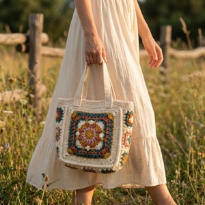May include: A cream-colored crochet tote bag with a colorful floral design. The bag features a square pattern with shades of teal, pink, yellow, and orange. The bag has two handles and is being held by a person wearing a cream-colored dress.