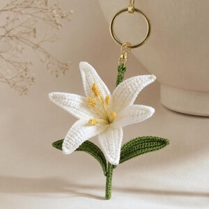 May include: A handmade crochet lily flower keychain. The white flower has yellow accents and green leaves and stem. The keychain has a gold-colored ring and clasp. The background is a soft, neutral color.