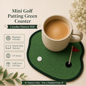 Puede incluir: Un posavasos de ganchillo verde con forma de mini campo de golf, con un borde negro, una bola de golf blanca y una bandera roja. Una taza beige llena de café se encuentra sobre el posavasos. El texto dice "Mini Golf Putting Green Coaster" y "Crochet Pattern PDF".