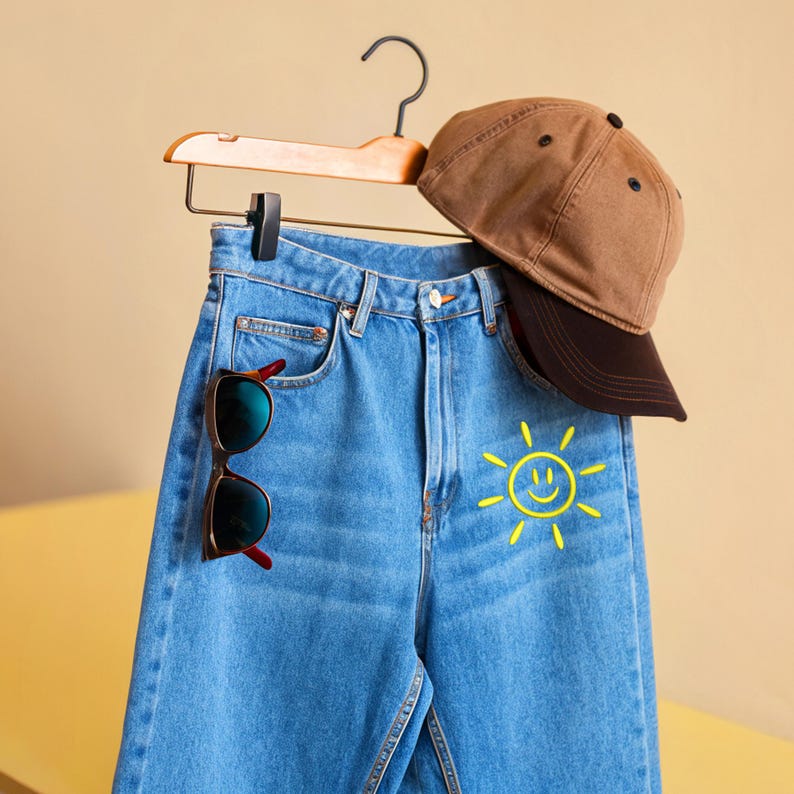 May include: Light blue denim jeans with a yellow smiling sun graphic, a brown baseball cap, and sunglasses with red accents hanging on a wooden hanger. The sunglasses have dark lenses.