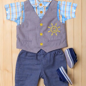 May include: A baby boy's outfit set on a wooden surface. The set includes a blue and orange plaid short-sleeved shirt, a gray vest with a yellow sun graphic, blue striped shorts, and navy blue shoes.