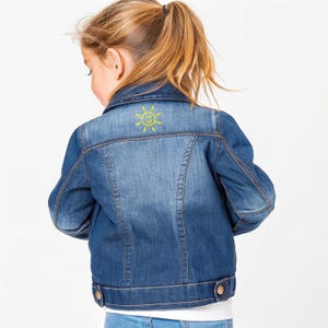 May include: A blue denim jacket with a yellow smiling sun design on the back. The jacket has a classic design with button closures and pockets. The denim is a medium wash, and the jacket appears to be for a child.