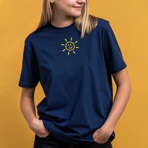 May include: Navy blue t-shirt featuring a yellow smiling sun graphic on the chest. The short-sleeved shirt is made of a soft material and is suitable for casual wear. The background is a solid yellow color.