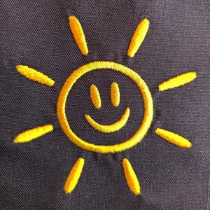 May include: Embroidered yellow sun design on a dark blue fabric. The sun features a smiling face and radiating lines, creating a cheerful and bright image. The embroidery has a textured appearance.