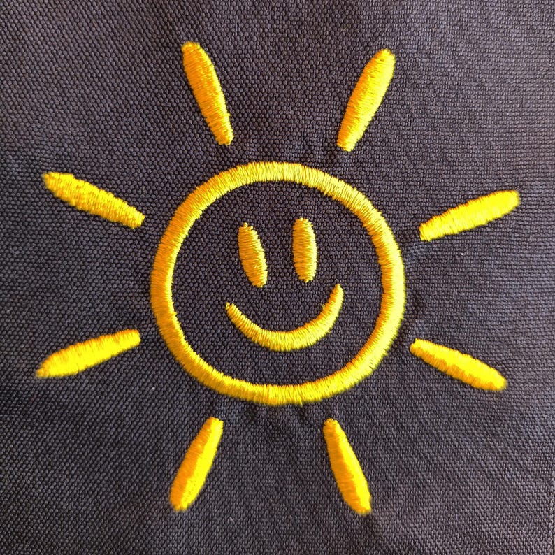 May include: Embroidered yellow sun design on a dark blue fabric. The sun features a smiling face and radiating lines, creating a cheerful and bright image. The embroidery has a textured appearance.