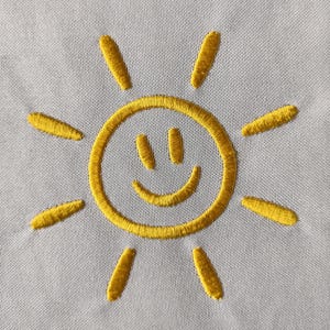 May include: Embroidered yellow sun design on a light gray fabric. The sun has a smiling face and radiating lines. The embroidery is detailed and the color is vibrant.