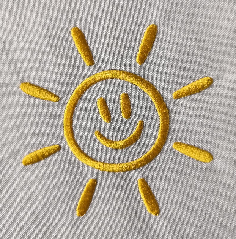 May include: Embroidered yellow sun design on a light gray fabric. The sun has a smiling face and radiating lines. The embroidery is detailed and the color is vibrant.