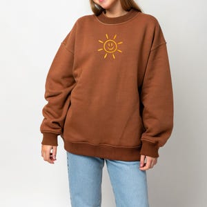 May include: A brown sweatshirt with a yellow smiling sun graphic. The sweatshirt has a crew neck, long sleeves, and ribbed cuffs and hem. The model is wearing light blue jeans.