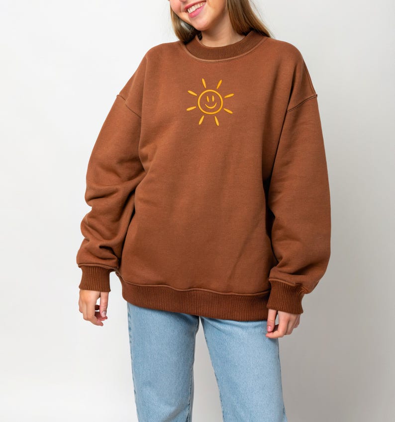 May include: A brown sweatshirt with a yellow smiling sun graphic. The sweatshirt has a crew neck, long sleeves, and ribbed cuffs and hem. The model is wearing light blue jeans.