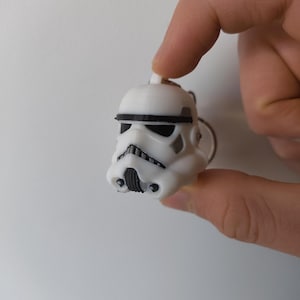 Może przedstawiać: Brelok w kształcie białego hełmu szturmowca z czarnymi detalami. Brelok jest miniaturową repliką kultowego hełmu z serii Gwiezdne Wojny. Hełm posiada czarne detale oczu i ust oraz czarny pasek na górze.