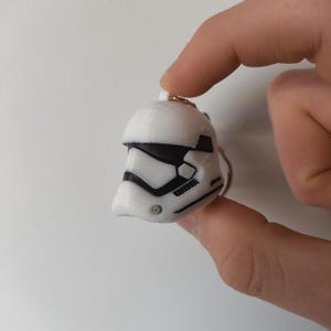 May include: A white and black Stormtrooper helmet keyring. The miniature helmet features the iconic design from the Star Wars franchise. The keyring is held by a silver ring.