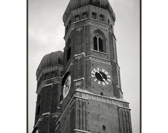 Puede incluir: Fotografía en blanco y negro de la Frauenkirche en Múnich, Alemania. La imagen captura las dos torres de la iglesia, cada una coronada por una cúpula distintiva. Las esferas del reloj son visibles en las torres y la mampostería es detallada.