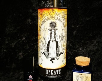 May include: A black glass candle with a yellow and orange label featuring a black and white illustration of a three-headed goddess. The label reads "HEKATE" and "rosariumblends.com". A small bottle of oil and a small jar of incense are also pictured, both with the same label.