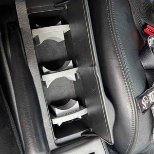 May include: A car's centre console with a built-in cup holder. The cup holder is grey and has three slots. The console is black and has a leather seat next to it.