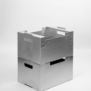 Puede incluir: Dos cajas de almacenamiento metálicas plateadas apiladas. Cada caja tiene forma rectangular con un recorte de asa en los laterales. Las cajas están remachadas en las esquinas y tienen un diseño minimalista.