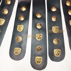May include: Close-up of black leather strips with gold-colored lion head and circular studs. The leather has a textured surface. The studs are evenly spaced along the strips, creating a decorative pattern. The image suggests a historical or fantasy theme.