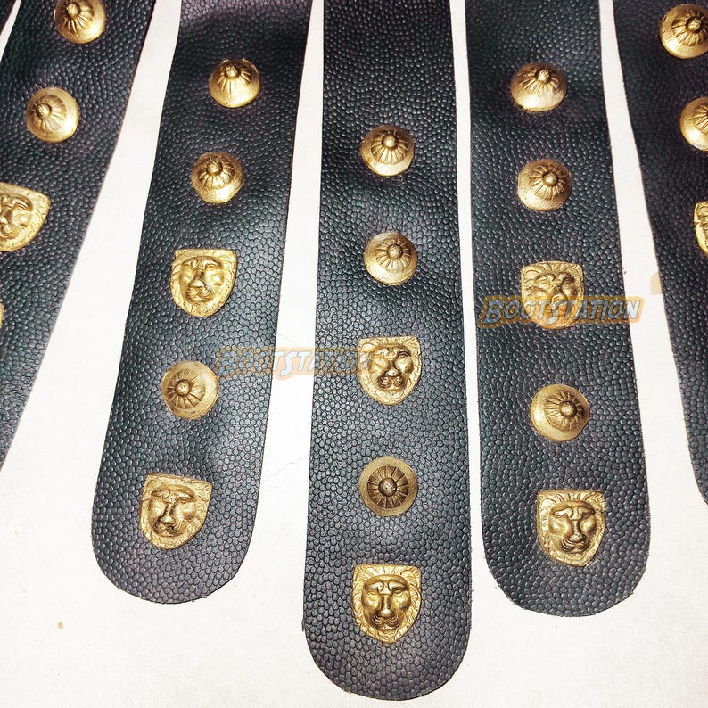 May include: Close-up of black leather strips with gold-colored lion head and circular studs. The leather has a textured surface. The studs are evenly spaced along the strips, creating a decorative pattern. The image suggests a historical or fantasy theme.