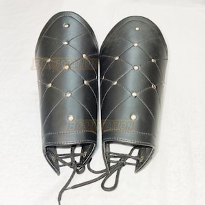 May include: A pair of black leather arm guards with diamond-shaped stitching and silver-colored studs. The arm guards have laces at the bottom and are designed to protect the forearms. The overall design suggests a medieval or historical theme.