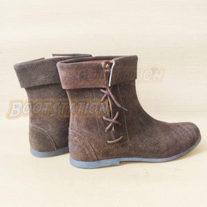 Könnte beinhalten: Ein Paar dunkelbraune Wildleder-Stiefeletten mit umgeschlagenem Schaft, abgerundeter Zehenpartie und seitlicher Schnürung mit dünnen braunen Schnürsenkeln. Die Stiefel haben eine flache Sohle und der Text BOOTSTATION ist oben sichtbar.