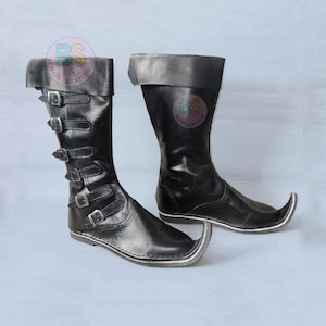 May include: A pair of black mid-calf boots featuring a pointed toe design and a folded cuff at the top. The left boot displays six horizontal straps with silver-toned metal buckles running up the side. A circular BS Bootstation logo is visible.