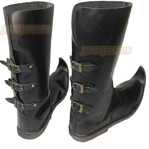 May include: A pair of black mid-calf boots featuring a pointed toe design and three vertical straps with brass-toned metal buckles along the back. The text BOOTSTATION is visible on each boot. The boots have a smooth finish and a flat sole.