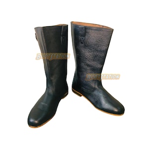 May include: A pair of black leather mid-calf boots with a textured finish, rounded toes, and flat soles. Each boot features a vertical stitched tab at the top edge and the text BOOTSTATION printed in orange and white across the side of the shaft.