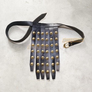 May include: Black leather belt with vertical strips, each adorned with brass-colored studs. The belt features a buckle and a loop for securing. The design suggests a historical or costume-related item.