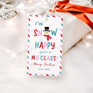 Puede incluir: Una etiqueta de regalo rectangular con una ilustración de muñeco de nieve y el texto "I'm SNOW HAPPY you're in MY CLASS! Merry Christmas From - Olivia". La etiqueta tiene un fondo blanco con un borde colorido y copos de nieve.