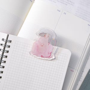 Op de afbeelding: Een transparante bladwijzer met een aquarel illustratie van een persoon in een roze jurk en een grijze hoed. De bladwijzer staat in een open notitieboek met gelinieerde pagina's, ideaal om een pagina te markeren.