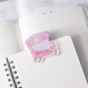Può includere: Un segnalibro trasparente in acrilico con un'illustrazione ad acquerello di una persona reclinata su una poltrona floreale rosa. Il segnalibro è posizionato sull'angolo di un quaderno aperto con un motivo a griglia.