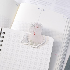 Può includere: Un segnalibro trasparente con un'illustrazione ad acquerello di una persona con cappotto e cappello bianchi, che tiene in mano un fascio. Il segnalibro è posto in un quaderno aperto con pagine a griglia, con la data 'mar 23' visibile.