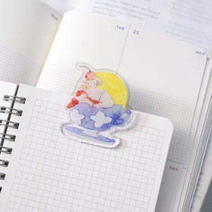 May include: A whimsical bookmark with a watercolour illustration of a person in a teacup against a yellow sun. The bookmark is in an open notebook with a grid pattern.