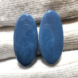 Könnte beinhalten: Zwei ovale, polierte Edelsteine in tiefblauer Farbe. Die Steine haben ein marmoriertes Aussehen mit helleren blauen Wirbeln. Sie liegen auf einem strukturierten, cremefarbenen Stoff.