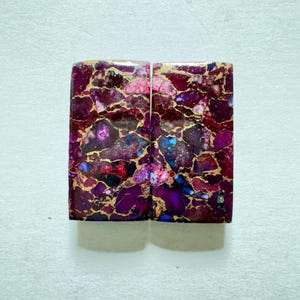 Puede incluir: Dos piedras rectangulares con un patrón multicolor vibrante. Las piedras presentan una mezcla de tonos morados profundos, rosas y azules, entrelazados con vetas doradas. Las superficies pulidas reflejan la luz, mostrando el intrincado diseño natural.