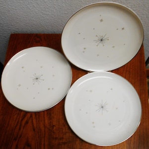 Op de afbeelding: Drie witte keramische borden met een gouden rand en een sterrenpatroon in het midden. De borden hebben een mid-century modern esthetiek, met kleine gouden steraccenten. De borden zijn gerangschikt op een donker houten oppervlak.