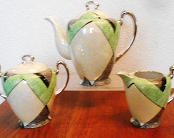 Puede incluir: Un juego de té vintage con tetera, azucarero y jarra de crema. El juego presenta una base de color crema con diseños geométricos verdes, negros y marrones. Las asas y los detalles plateados completan el conjunto. El juego se exhibe sobre una superficie de madera.