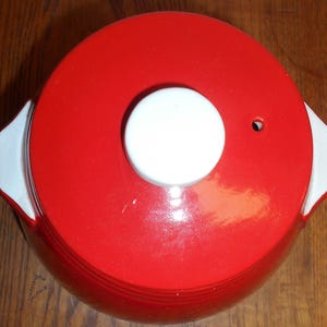 May include: A red ceramic casserole dish with a white handle on each side. The lid is red with a white knob and a small hole. The dish is on a wooden surface.