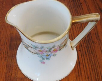 May include: A white porcelain creamer with a gold rim and floral design. The creamer has a flared base and a handle with a gold trim.
