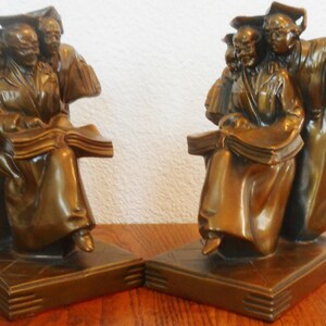 Puede incluir: Un par de sujetalibros de bronce que representan a un grupo de personas con batas de graduación sosteniendo libros. Los sujetalibros están sobre una superficie de madera.
