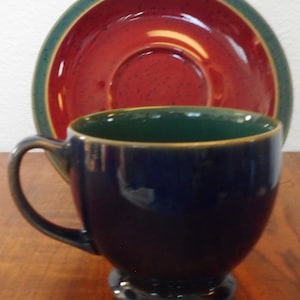 May include: A blue and green ceramic teacup and saucer set. The saucer has a red centre with green and gold trim. The teacup has a blue body with a green rim.