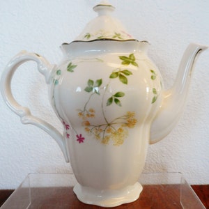 Puede incluir: Tetera de porcelana blanca con un diseño floral con flores rosas, amarillas y verdes. La tetera tiene una tapa y un pico.
