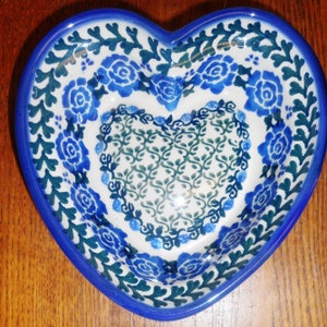 May include: Heart-shaped ceramic bowl with a blue rim and floral patterns. The bowl features a white interior with blue rose and leaf designs. The outer edge has a dark blue border. The bowl is approximately 8 inches wide.
