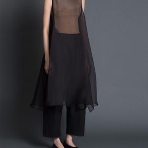 Puede incluir: Una túnica negra sin mangas sobre una camiseta y pantalones negros. La túnica es transparente, con cuello alto. Los pantalones son cortos y los zapatos son bailarinas negras. El fondo es gris liso.