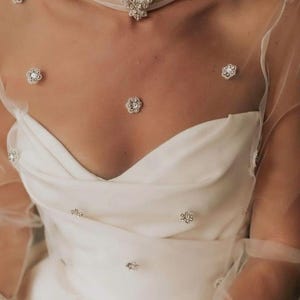 Op de afbeelding: Een witte trouwjurk met een sweetheart-halslijn en een doorschijnende overlay versierd met sprankelende bloemversieringen. Een bijpassende choker-ketting met een centraal juweel en bloemaccenten maakt de elegante look compleet.