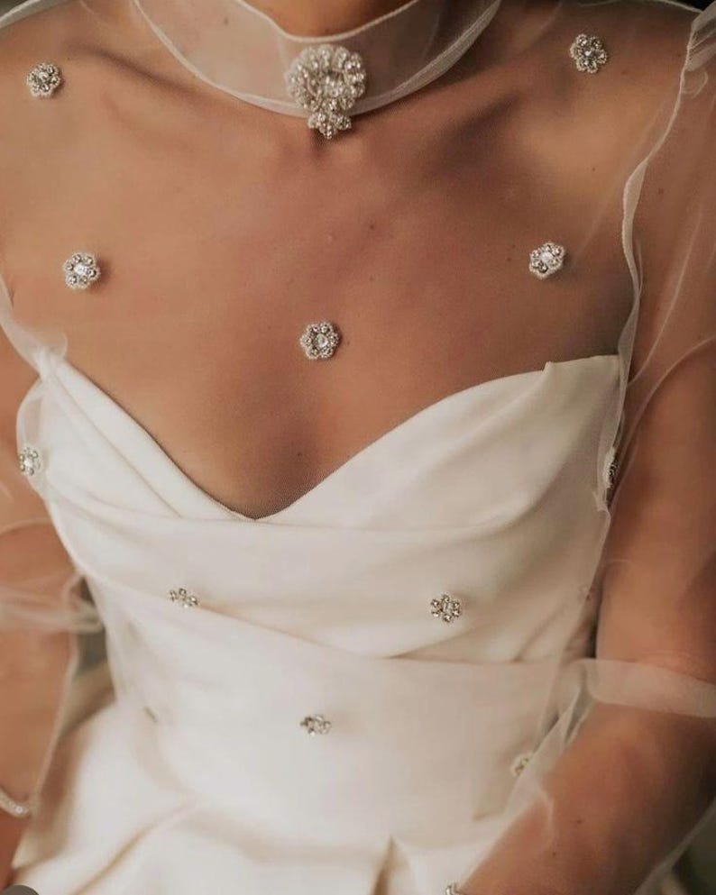 Op de afbeelding: Een witte trouwjurk met een sweetheart-halslijn en een doorschijnende overlay versierd met sprankelende bloemversieringen. Een bijpassende choker-ketting met een centraal juweel en bloemaccenten maakt de elegante look compleet.